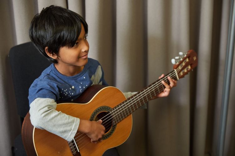 A boy with black hair and long sleeved blue and grey top smiles away from the camera as he plays the guitar